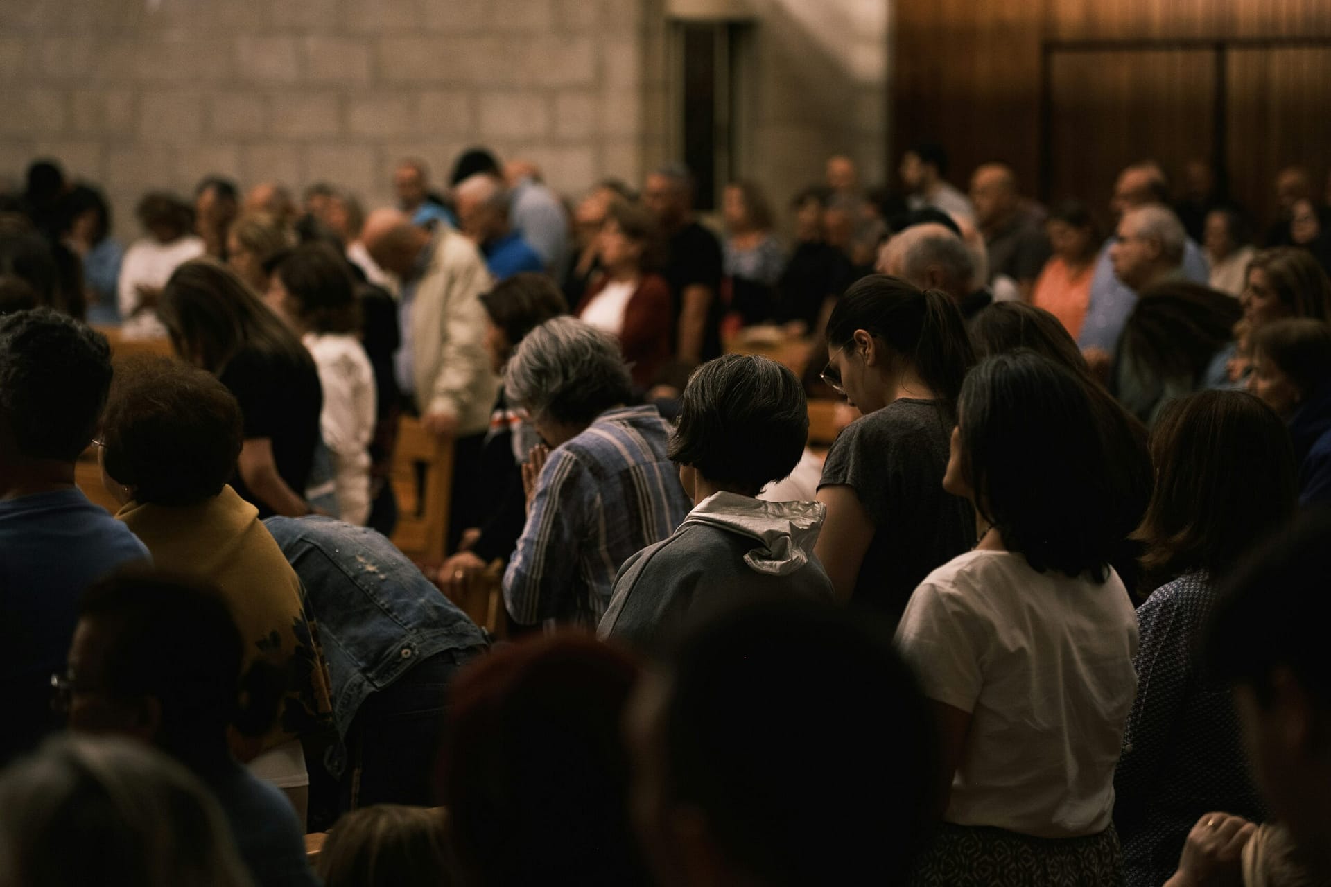A large group of people stands closely together in a dimly lit indoor setting, suggesting a solemn or reflective event. They are facing forward, some with heads bowed.